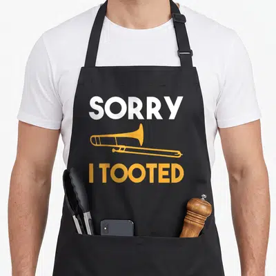 Sorry trombone quotes apron - Orange trombone graphic between white SORRY and orange I TOOTED text — shown on a black cotton canvas apron against a white background