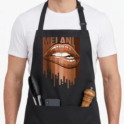 Melanin magic apron - MELANIN text above glossy brown lips over vertical dripping stripes in cartoon style — shown on a black cotton canvas apron against a white background