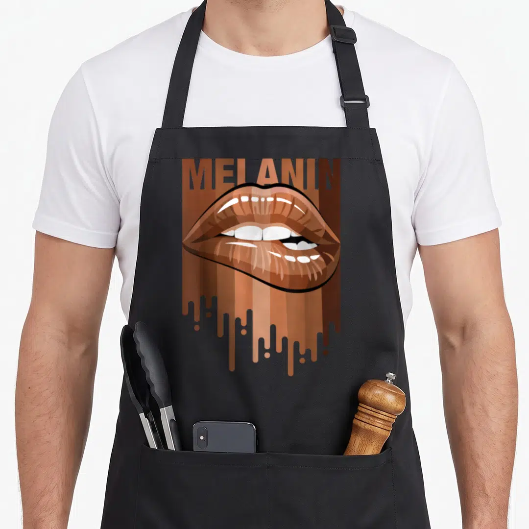Melanin magic apron - MELANIN text above glossy brown lips over vertical dripping stripes in cartoon style — shown on a black cotton canvas apron against a white background