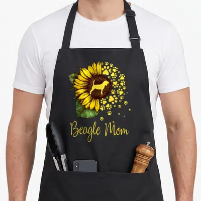 Sunflower beagle apron - Beagle Mom text below sunflower with dog silhouette and scattered yellow paw prints — shown on a black cotton canvas apron against a white background