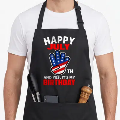 Funny birthday 4th of july apron - Distressed hand filled with US flag pattern surrounded by Happy July Th And Yes — shown on a black cotton canvas apron against a white background
