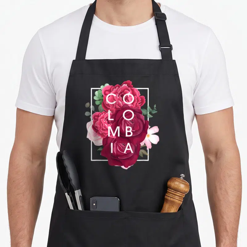 Flower colombia pride apron - Vertical COLOMBIA text overlaid on red and pink roses inside a white rectangle — shown on a black cotton canvas apron against a white background