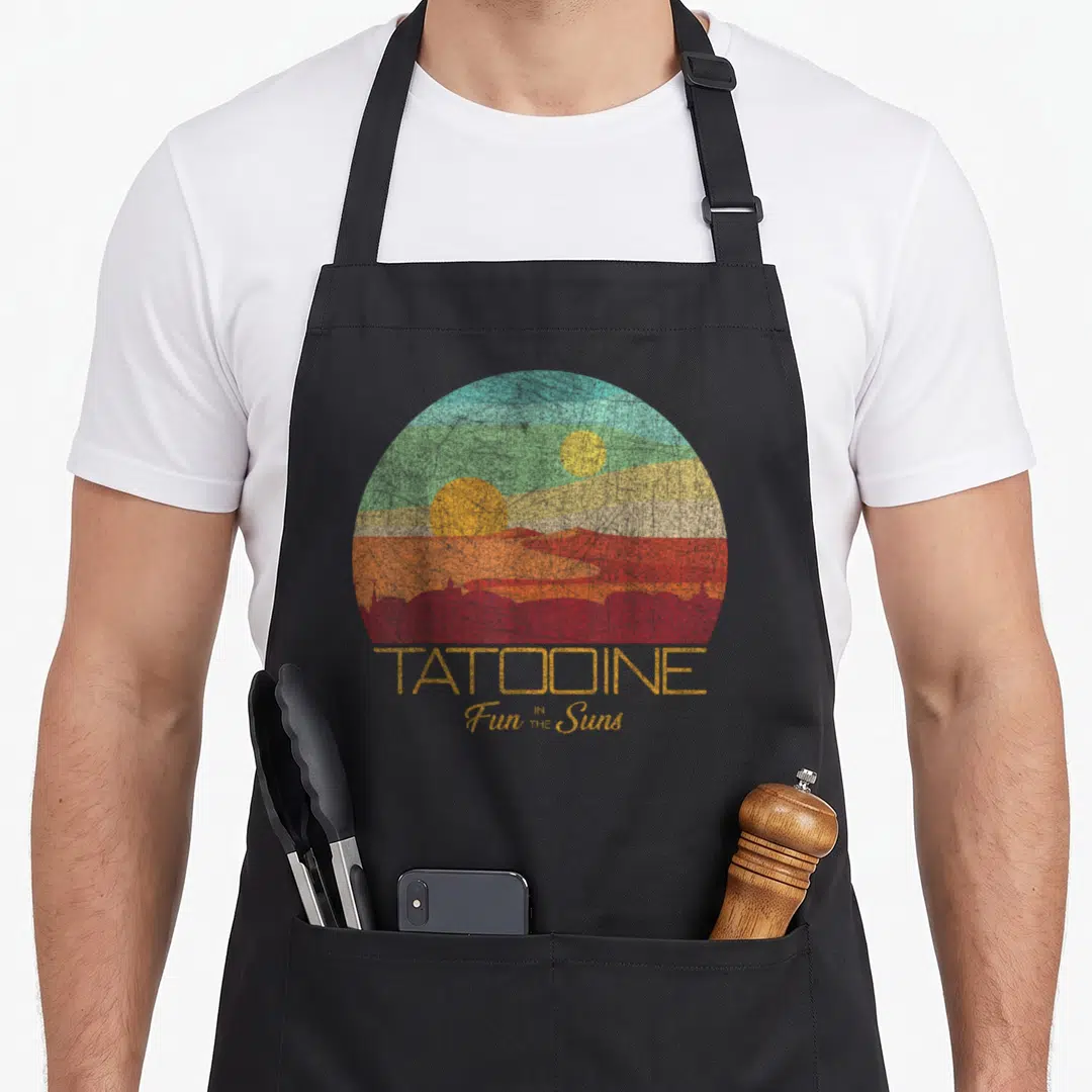 Postcard star war apron - Distressed semi-circle landscape with two suns, dunes, skyline, and TATOOINE Fun IN THE Suns — shown on a black cotton canvas apron against a white background