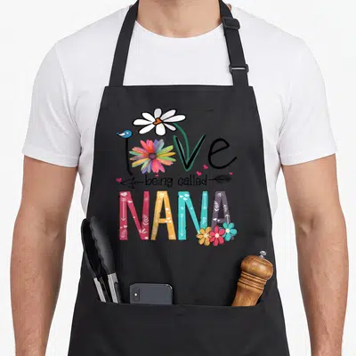 Nana floral aprons - Love being called NANA text art with rainbow flower, blue bird, and patterned colorful block — shown on a black cotton canvas apron against a white background