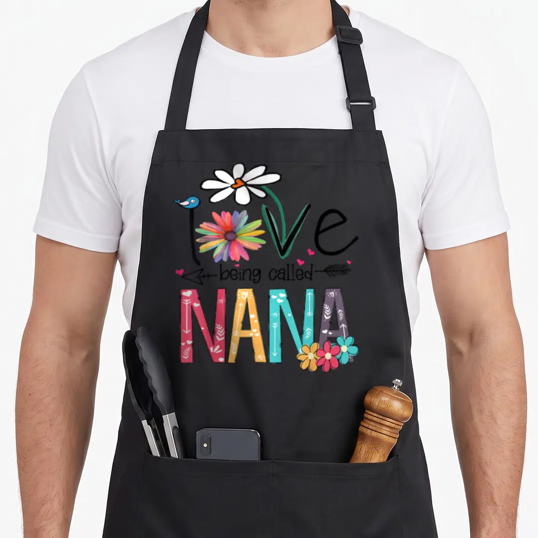 Nana floral aprons - Love being called NANA text art with rainbow flower, blue bird, and patterned colorful block — shown on a black cotton canvas apron against a white background