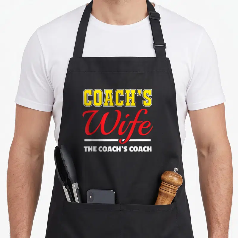Coaching wife apron - COACH'S Wife THE COACH'S COACH text in yellow red and white typography — shown on a black cotton canvas apron against a white background