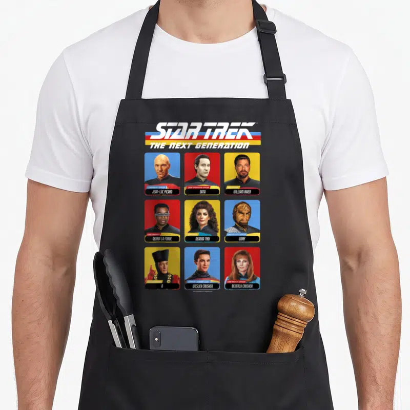 Cast star apron - Nine Star Trek TNG character portraits in grid with role labels and names — shown on a black cotton canvas apron against a white background