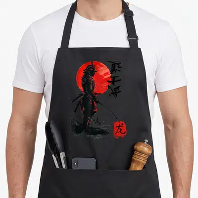 Samurai eclipse apron - Black samurai silhouette against red sun with vertical calligraphy and red seal box — shown on a black cotton canvas apron against a white background