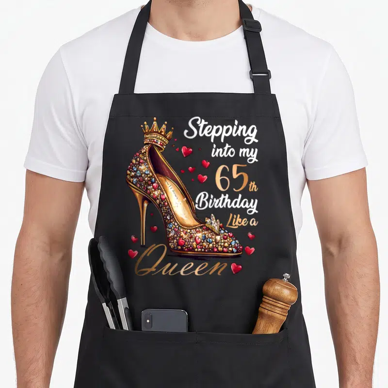 Stepping 65th queen birthday apron - Gold jeweled high heel shoe with crown and red hearts next to birthday text — shown on a black cotton canvas apron against a white background