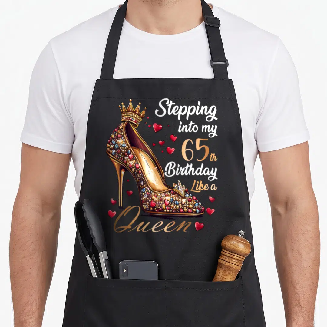 Stepping 65th queen birthday apron - Gold jeweled high heel shoe with crown and red hearts next to birthday text — shown on a black cotton canvas apron against a white background