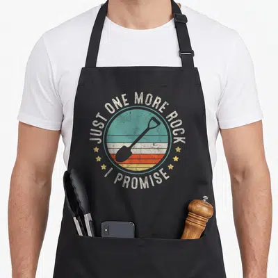 Geology geologist apron - Grey shovel on teal orange yellow beige striped circle with arched text and three stars each — shown on a black cotton canvas apron against a white background