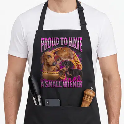 Proud dachshund apron - Dachshunds and hot dogs between PROUD TO HAVE A SMALL WIENER text with purple lightning — shown on a black cotton canvas apron against a white background