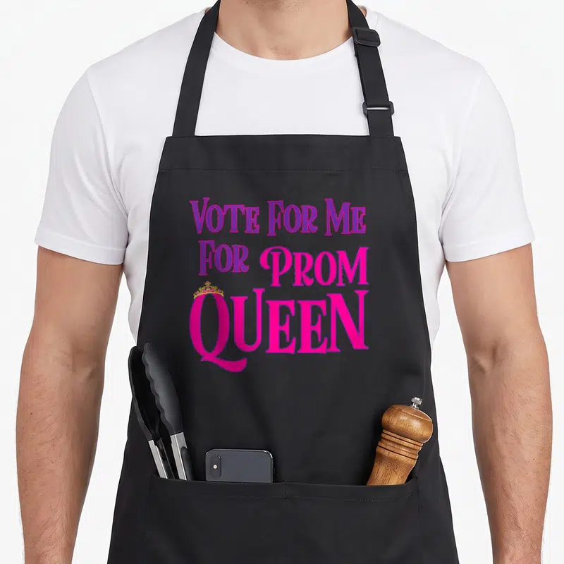 Vote queen apron - Stacked purple and pink text reading VOTE FOR ME FOR PROM QUEEN with a small crown on the Q — shown on a black cotton canvas apron against a white background