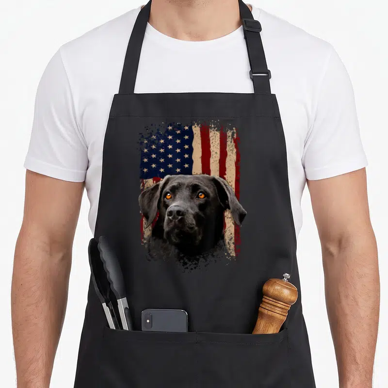 Labrador american flag apron - Realistic black dog head centered on distressed red white and blue flag background — shown on a black cotton canvas apron against a white background