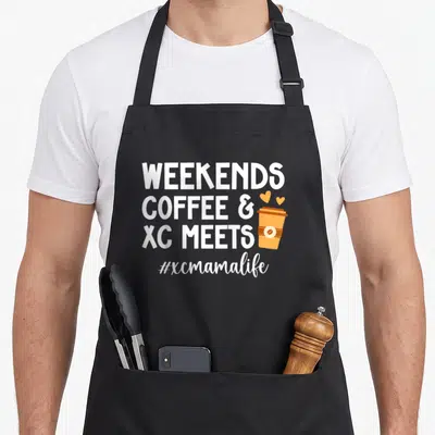 Mama cross country apron - Stacked white text WEEKENDS COFFEE XC MEETS next to orange coffee cup with two hearts — shown on a black cotton canvas apron against a white background