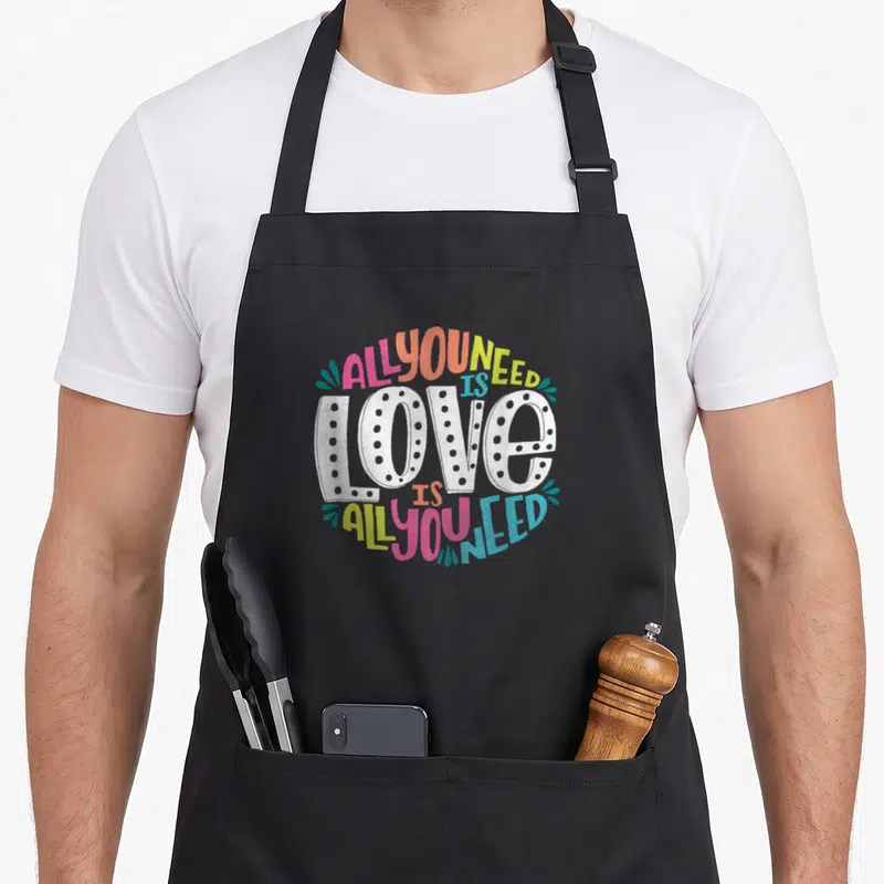Product valentine's day apron - Circular text design reading ALL YOU NEED IS LOVE in pink orange yellow blue and white — shown on a black cotton canvas apron against a white background