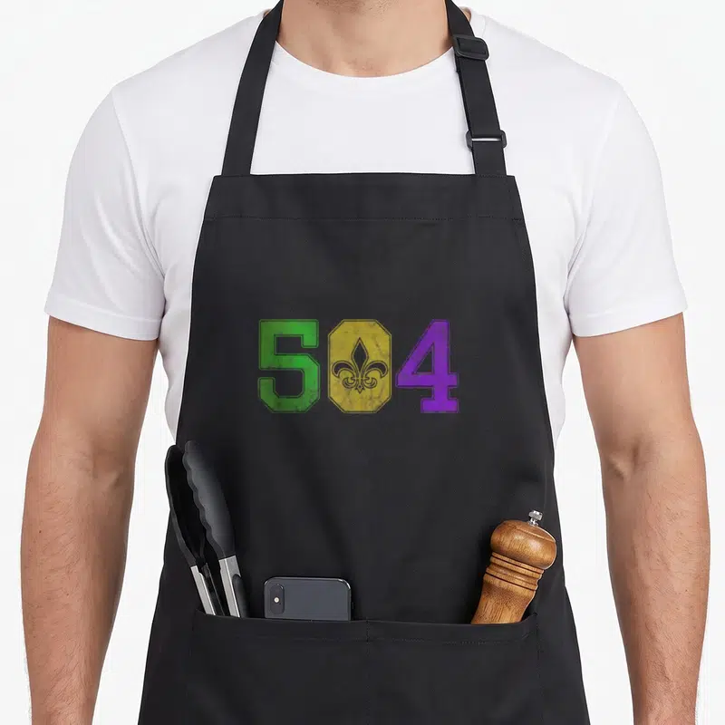 Mardi louisiana apron - Green 5 and purple 4 flanking a yellow shield with a purple fleur-de-lis representing the 504 — shown on a black cotton canvas apron against a white background