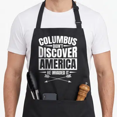 Aboriginal native american apron - White stacked text Columbus didn't discover America he invaded it above two crossed — shown on a black cotton canvas apron against a white background