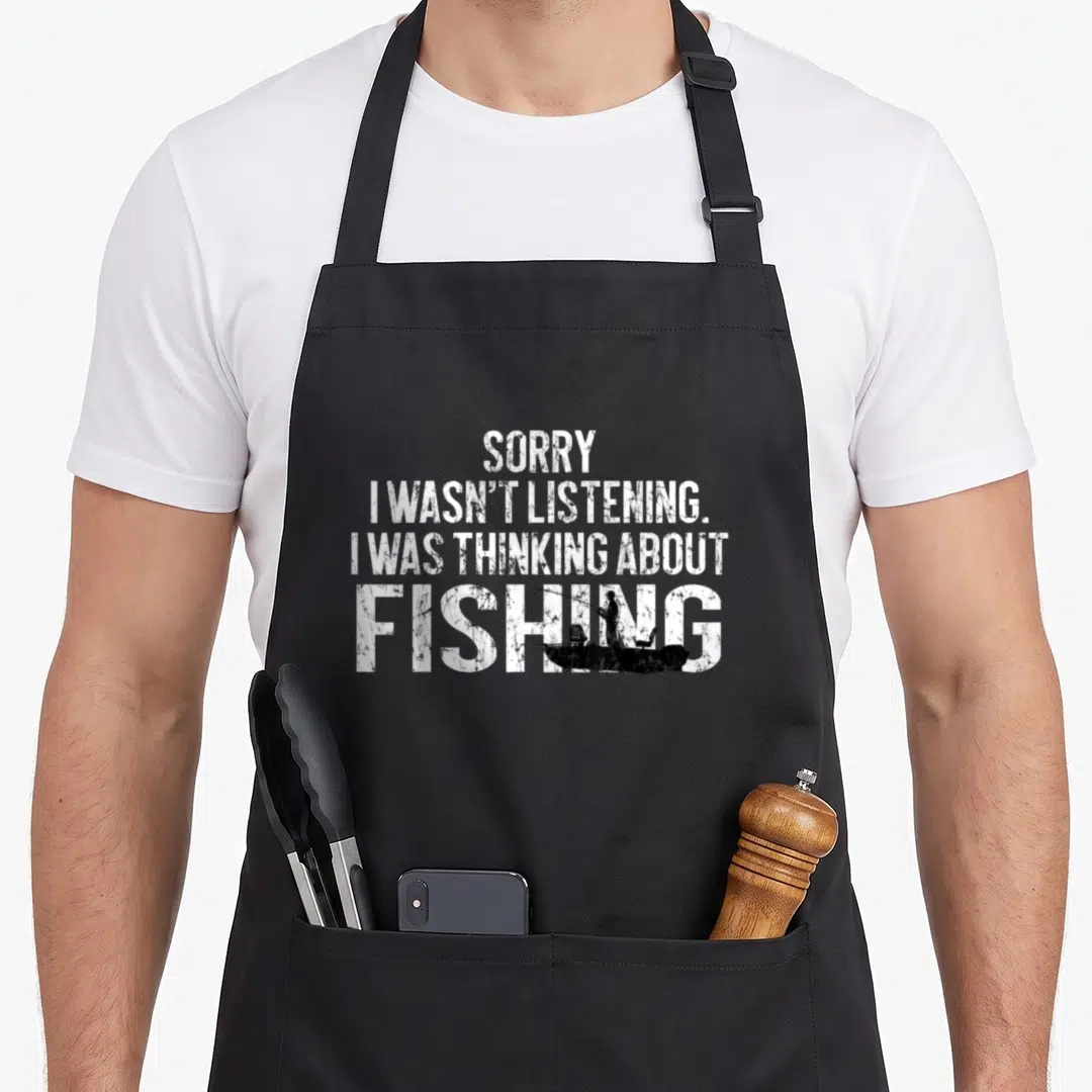 Fishing joke apron - White distressed text Sorry I Wasnt Listening with fisherman silhouette in boat over Fishing — shown on a black cotton canvas apron against a white background