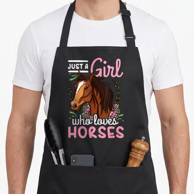 Jumping horse apron - Cartoon brown horse head with pink flowers and text Just a Girl who loves Horses — shown on a black cotton canvas apron against a white background