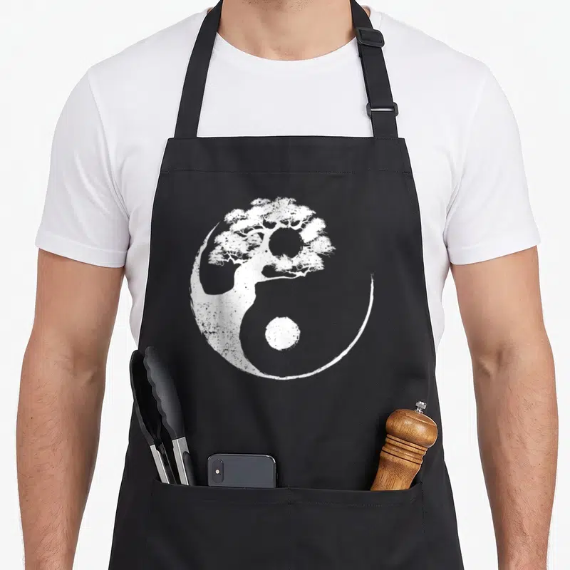 Yang buddhist tree apron - White yin yang symbol with integrated tree on black apron — shown on a black cotton canvas apron against a white background