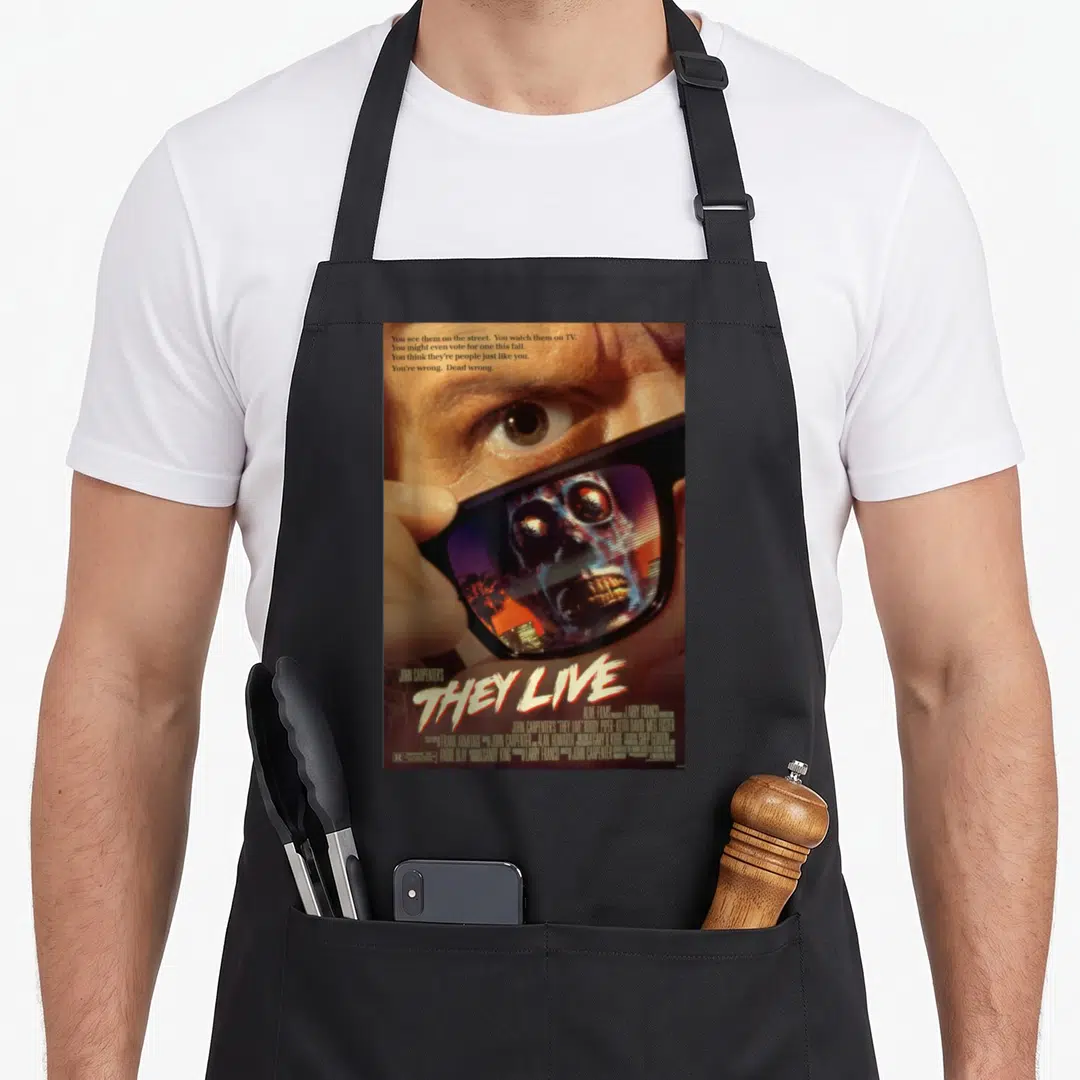 Live they think apron - Man holding sunglasses revealing alien skull face with They Live text in brown black blue white — shown on a black cotton canvas apron against a white background
