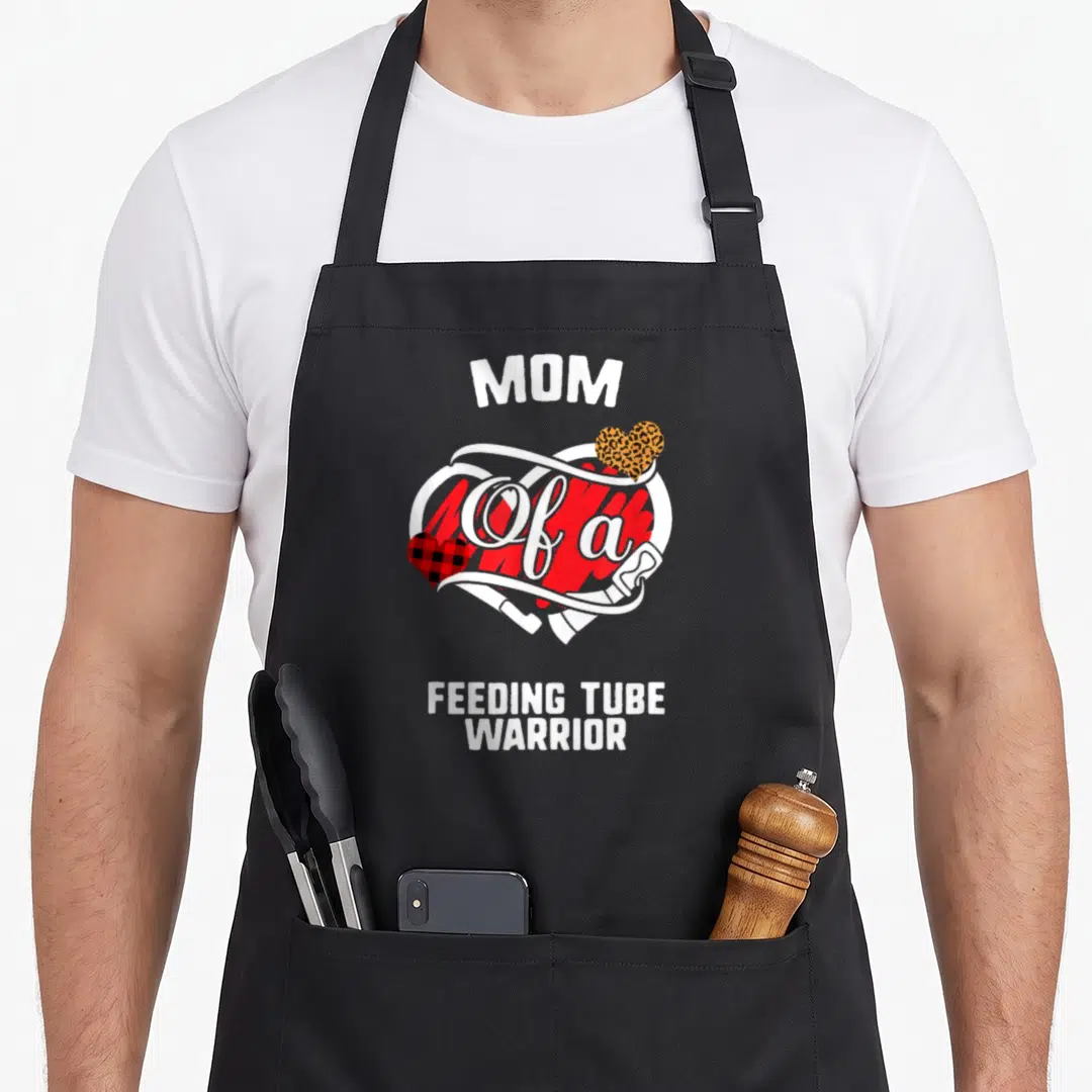 Feeding buffalo plaid apron - MOM Of a FEEDING TUBE WARRIOR text with red heart, white tube, and patterned hearts — shown on a black cotton canvas apron against a white background