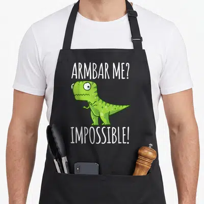 Bjj t-rex apron - Green cartoon dinosaur with white text reading armbar me impossible above and below — shown on a black cotton canvas apron against a white background