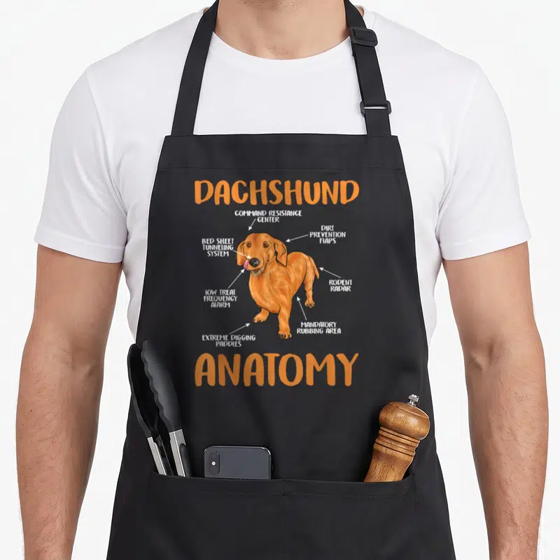 Anatomy wiener apron - Cartoon dachshund labeled with humorous anatomy terms like Rodent Radar and Extreme Digging — shown on a black cotton canvas apron against a white background