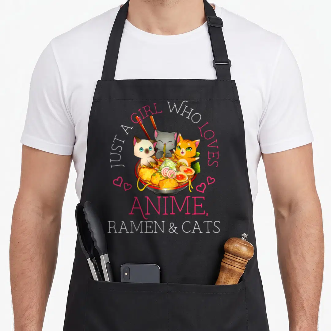Ramen manga cat apron - Three cartoon cats inside a ramen bowl with arched pink and white text — shown on a black cotton canvas apron against a white background