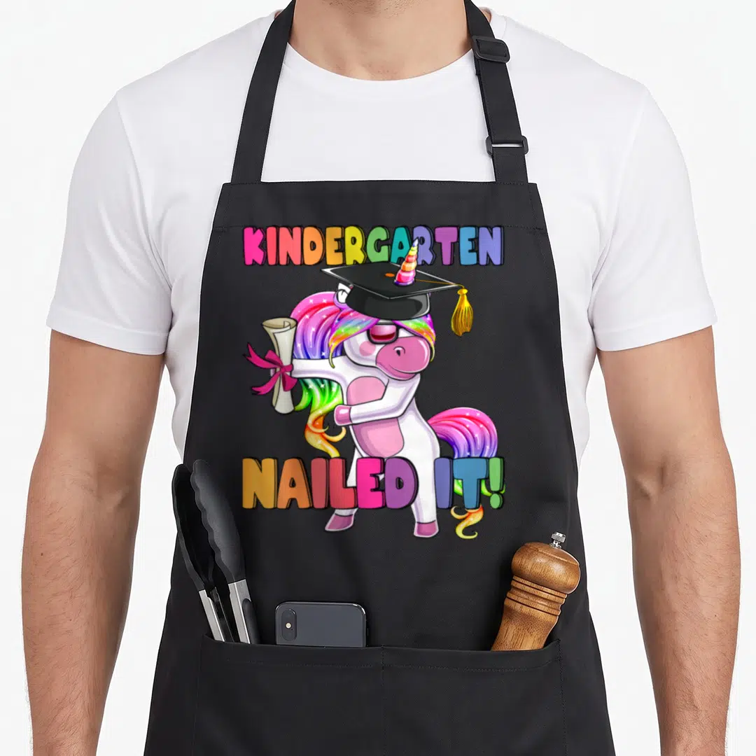 Kindergarten flossing unicorn aprons - Rainbow text Kindergarten Nailed It with flossing unicorn in graduation cap — shown on a black cotton canvas apron against a white background