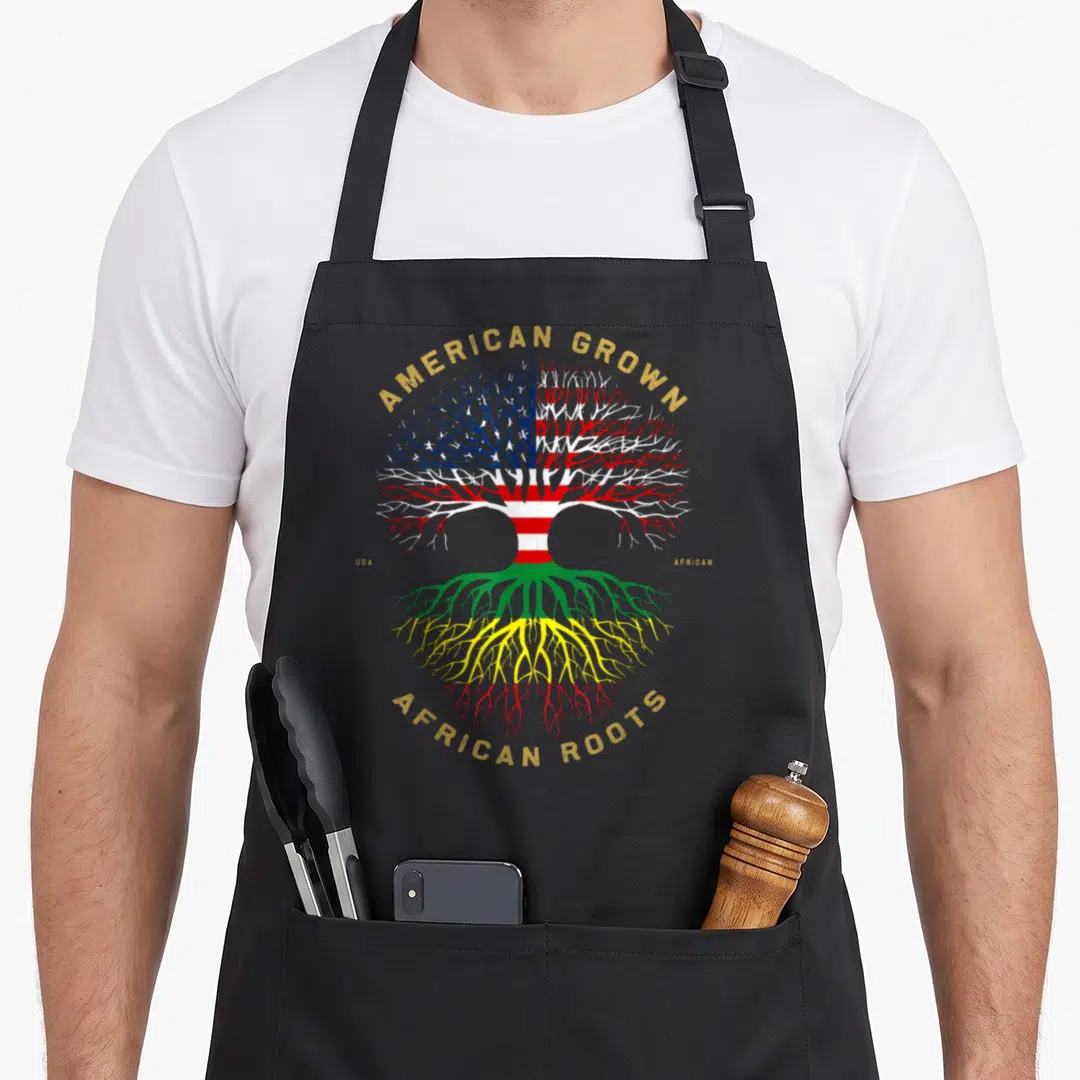 African american flag apron - Tree with US flag branches and green yellow roots surrounded by gold text American Grown — shown on a black cotton canvas apron against a white background