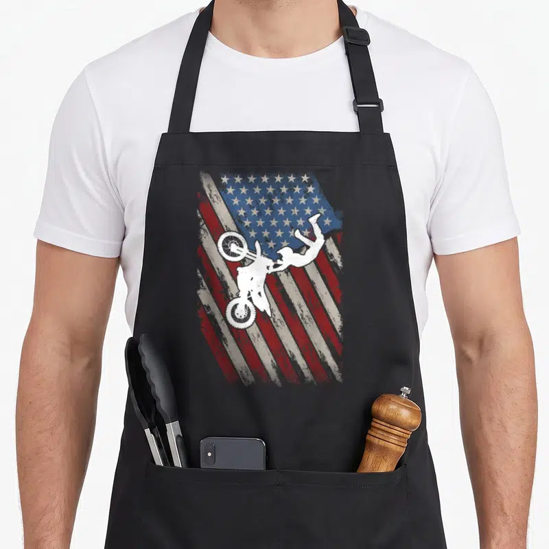 Bike 4th of july apron - White motocross rider silhouette on distressed red white and blue flag — shown on a black cotton canvas apron against a white background