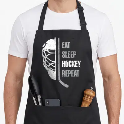 Eat christmas apron - White distressed hockey mask and stick beside stacked EAT SLEEP HOCKEY REPEAT text — shown on a black cotton canvas apron against a white background
