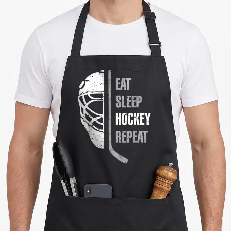 Eat christmas apron - White distressed hockey mask and stick beside stacked EAT SLEEP HOCKEY REPEAT text — shown on a black cotton canvas apron against a white background