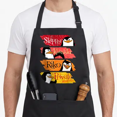 Panel penguin aprons - Four stacked banners with penguin faces and names Skipper Kowalski Riko Private in red orange — shown on a black cotton canvas apron against a white background