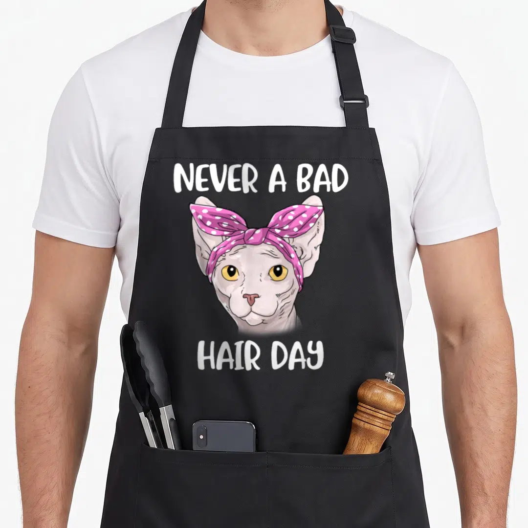 Vintage sphynx cat apron - Cartoon sphynx cat head with pink polka dot bow and white text reading NEVER A BAD HAIR DAY — shown on a black cotton canvas apron against a white background