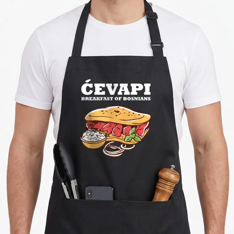 Cevapi food aprons - Cartoon ćevapi sandwich with ĆEVAPI BREAKFAST OF BOSNIANS text, dip bowl, and onion rings in white — shown on a black cotton canvas apron against a white background
