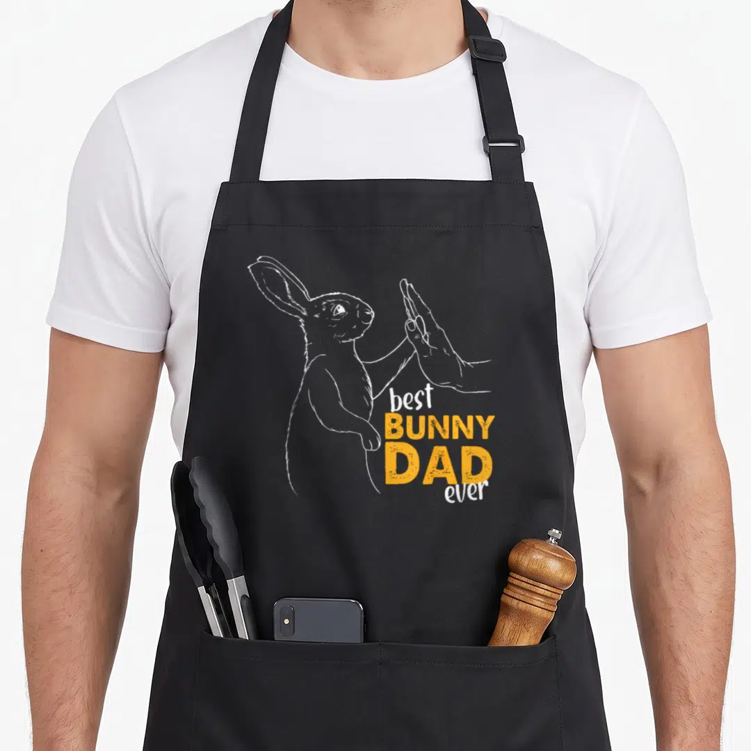 Bunny daddy apron - White line-art bunny high-fiving human hand with stacked orange and white text — shown on a black cotton canvas apron against a white background
