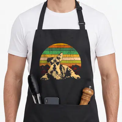 Medium schnauzer apron - Retro dog portrait in front of orange yellow green striped sun with two red tree silhouettes — shown on a black cotton canvas apron against a white background