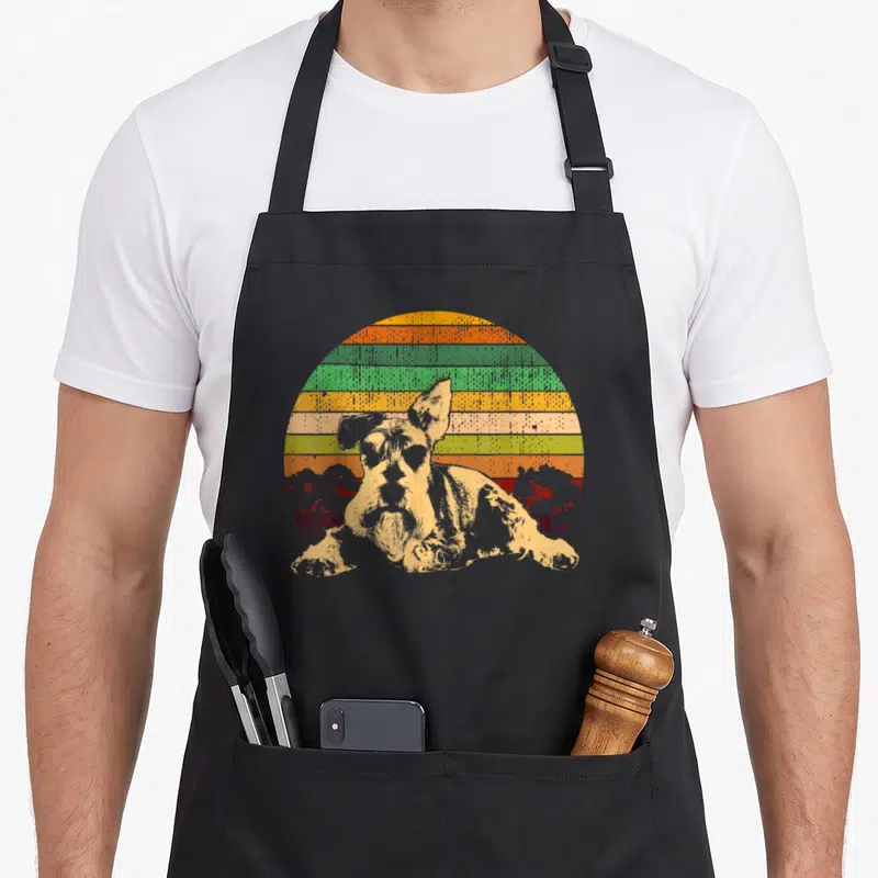 Medium schnauzer apron - Retro dog portrait in front of orange yellow green striped sun with two red tree silhouettes — shown on a black cotton canvas apron against a white background