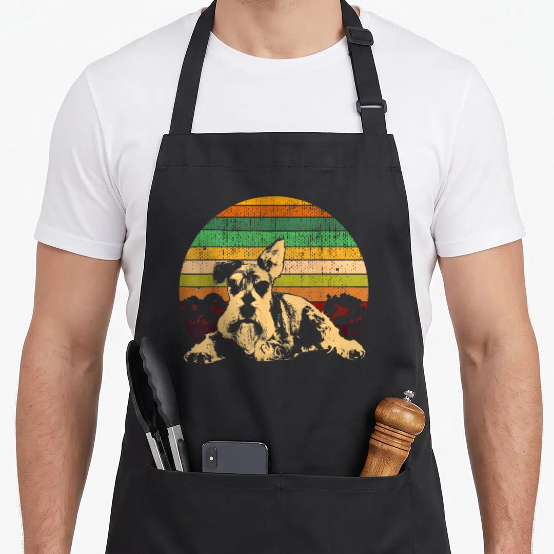 Medium schnauzer apron - Retro dog portrait in front of orange yellow green striped sun with two red tree silhouettes — shown on a black cotton canvas apron against a white background