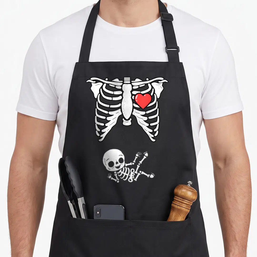 Announcement halloween apron - Cartoon white ribcage with red heart above a baby skeleton lying on its back — shown on a black cotton canvas apron against a white background