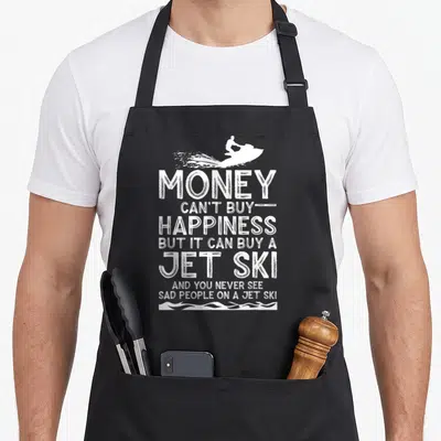 Ski jetski apron - White distressed text reading Money Can't Buy Happiness But It Can Buy A Jet Ski under a rider — shown on a black cotton canvas apron against a white background