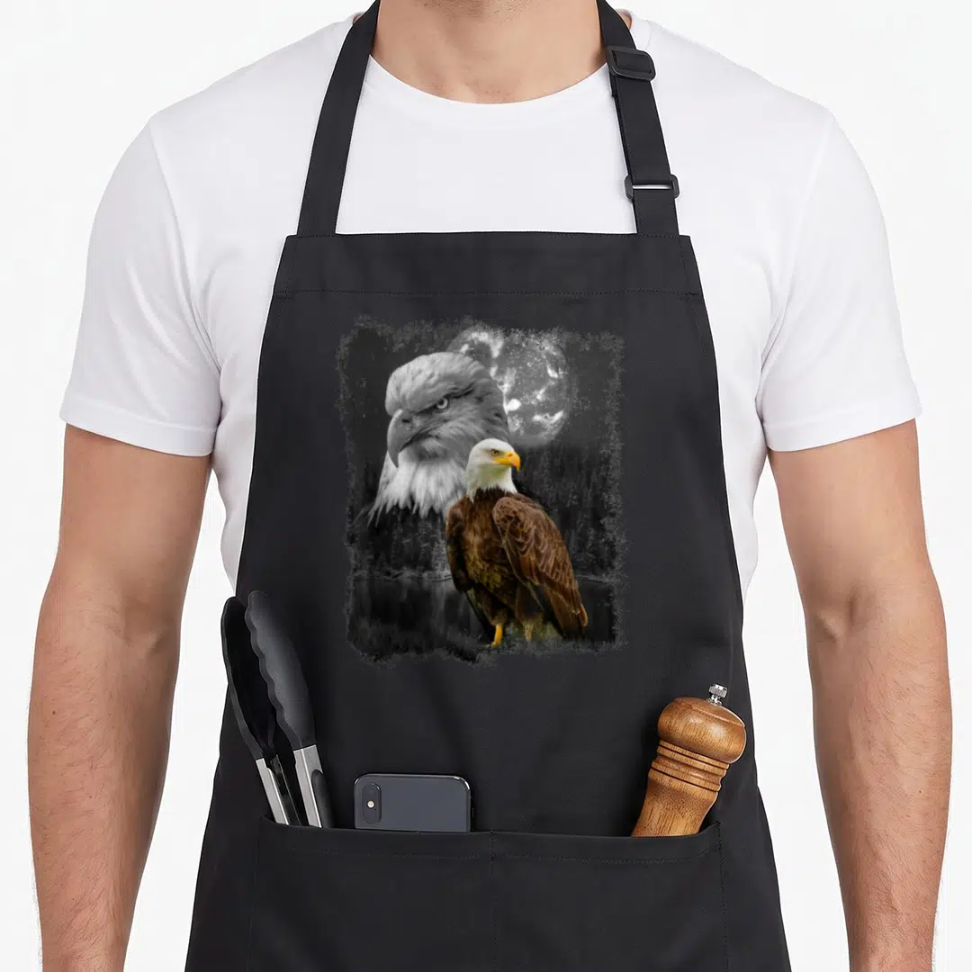 Nature eagles apron - Full-body bald eagle standing before a large moon head inside a rough-edged square frame — shown on a black cotton canvas apron against a white background