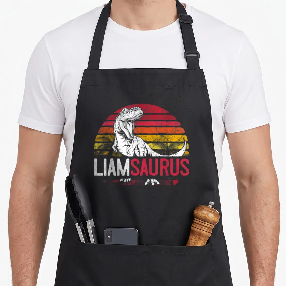 Liam dinosaur apron - White T-Rex over red orange yellow striped sun with LIAMSAURUS text — shown on a black cotton canvas apron against a white background