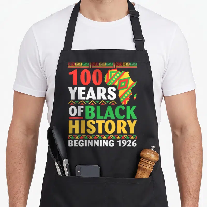 History centennial apron - 100 years of black history beginning 1926 text with patterned africa map and geometric — shown on a black cotton canvas apron against a white background