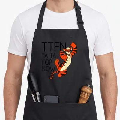 Tiger novelty apron - Cartoon tiger jumping next to stacked TTFN ta ta for now text in orange and black — shown on a black cotton canvas apron against a white background
