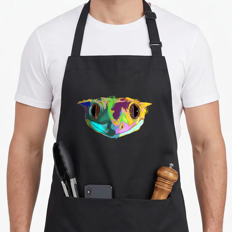 Reptile colourful apron - Cartoon gecko head in yellow, purple, green, blue, and pink — shown on a black cotton canvas apron against a white background