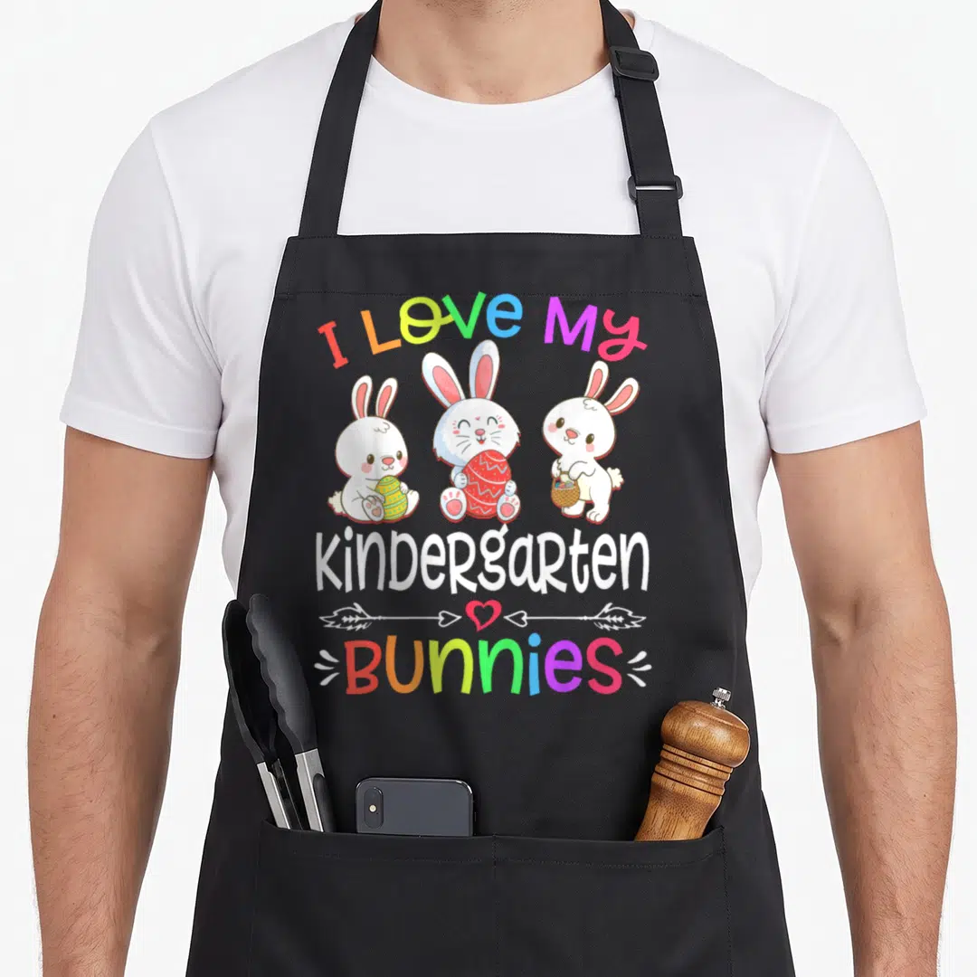 Bunny kindergarten teacher apron - Three cartoon bunnies holding eggs with I Love My Kindergarten Bunnies rainbow text — shown on a black cotton canvas apron against a white background
